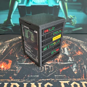 May include: A black, rectangular server rack miniature featuring a textured finish. The front side displays a vertical card slot containing a card labeled Firestarter. The side panel features five horizontal server modules with red and green lights.