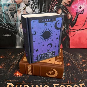 May include: A vertical blue book-shaped storage box with a sun, moon, and star pattern and the word MYSTICS on the front, resting on a horizontal brown book-shaped storage box with a moon and star icon and the word MYSTICS on the spine.