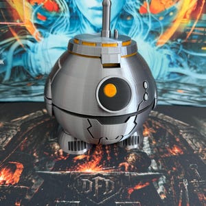 May include: A spherical silver droid figure with a central black circular eye featuring a bright orange center. The droid has a small vertical antenna on top, a horizontal orange band around the upper section, and two small feet at the base.
