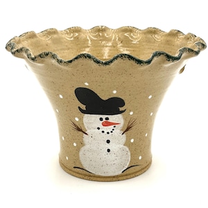 May include: A beige ceramic vase with a snowman design. The vase has a ruffled rim with a green trim. The snowman has a black hat, a carrot nose, and twig arms. White dots are scattered around the snowman.