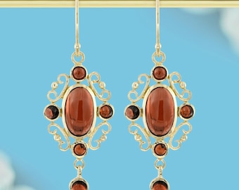 May include: A pair of gold-colored earrings with an ornate design. Each earring features a large, oval-shaped, reddish-brown stone at the center, surrounded by smaller, circular stones of a similar color. The earrings hang from a gold-colored hook.