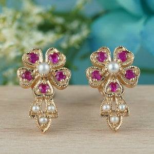 May include: A pair of gold earrings with a flower design. Each earring has five pink gemstones and a single pearl in the center.