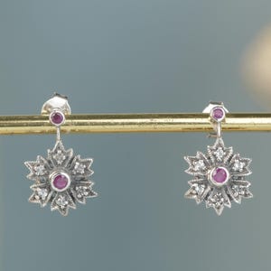 May include: A pair of silver drop earrings featuring a central round ruby surrounded by small clear stones in a star-shaped setting. Each earring has a smaller round ruby accent at the top post, creating a symmetrical floral design.