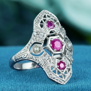 May include: A silver-toned ring with an ornate, Art Deco design. The ring features three vibrant, round, ruby-colored gemstones and several smaller clear stones. Two small pearls are also incorporated into the design. The ring is set against a blue background.