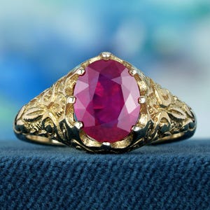 May include: A gold ring featuring an oval ruby gemstone set in an intricate, ornate design.