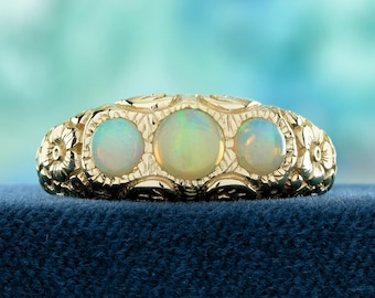 May include: A gold ring featuring three oval opals. The ring has a floral design on the band and around the opals. The opals have a milky, iridescent appearance. The ring is set against a blue background.