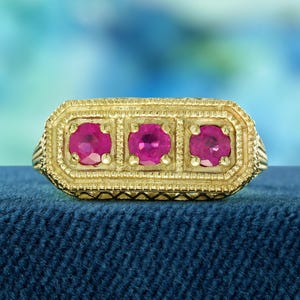 May include: A gold ring with three ruby stones set in a rectangular frame. The ring has a detailed design with a textured surface.