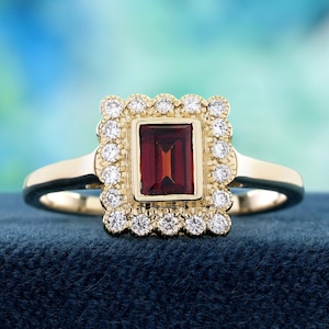 May include: A gold ring with a rectangular, deep red gemstone at its centre, framed by small, round, clear gemstones. The ring's band is plain, polished gold. The setting is on a dark blue surface.