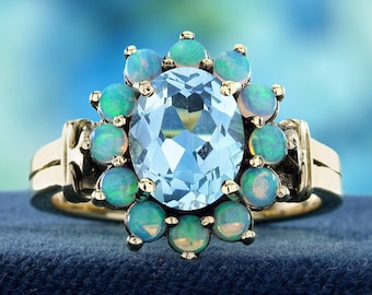 May include: A gold ring featuring a large, oval-cut light blue gemstone at the center, surrounded by a halo of ten small, round, iridescent opals. The band is made of polished yellow gold, providing a bright contrast to the blue and opal stones.
