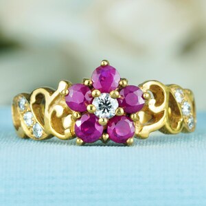 Könnte beinhalten: Ein goldener Ring mit floralem Design. Er hat sechs oval geschliffene, tiefrosa Rubine, die einen zentralen, runden, klaren Diamanten umgeben. Das Band hat ein dekoratives Filigranmuster mit kleinen, klaren Akzentsteinen.