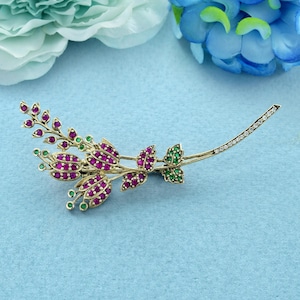 Puede incluir: Un broche floral dorado con piedras preciosas de color rubí y verde. El broche tiene un tallo curvo con una línea de pequeñas piedras transparentes en el extremo. El diseño incluye racimos de piedras de color rubí y detalles verdes.