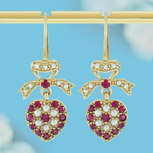 May include: A pair of gold-toned drop earrings. Each earring features a bow design with small white stones and a central red stone. Below, a heart-shaped pendant is adorned with red and white stones.
