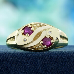 May include: A gold ring with two oval ruby gemstones and small diamond accents. The ring has a unique design with a flowing, organic shape. The ring is set against a blue background.