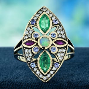 Op de afbeelding: Een gouden ring met een sierlijk ontwerp met een centrale opaal, twee smaragden, twee robijnen en verschillende kleine blauwe edelstenen. De ring is verder versierd met talrijke kleine diamanten, wat een luxueus en gedetailleerd sieraad creëert.
