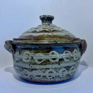May include: A ceramic lidded pot with a rustic design. The pot is primarily a light blue-gray color with brown accents and a white, chain-like pattern. It has two handles and a knob on the lid.
