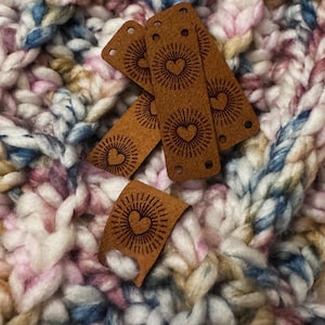 May include: Four brown leather labels with a heart and sunburst design. The labels are on a white, pink, and blue crocheted surface.
