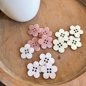 May include: Assortment of pink and white flower-shaped buttons with five petals and four holes in the center. The buttons are arranged on a wooden surface.