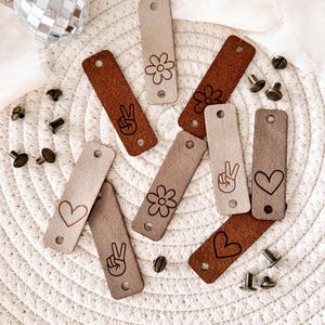 May include: Eight leather tags with different designs, including peace signs, hearts, and flowers. The tags are brown and beige and have holes punched in them.
