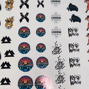 Puede incluir: Una hoja de pegatinas con varios diseños y logotipos. Los diseños incluyen X's negros, formas geométricas, las palabras "LOST LANDS", "LSDREAM" y "KAI WACHI", junto con otros gráficos y texto. Las pegatinas son en negro, blanco, dorado y azul.