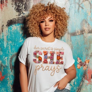 May include: White t-shirt with the text "Her secret is simple SHE prays" in a floral design. The word "SHE" is filled with colorful flowers. The shirt has short sleeves and a relaxed fit.