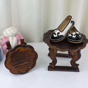 May include: A pair of white and black flats with a decorative design, resting on a small, dark wooden table. The shoes have a rounded toe and a contrasting black trim. A second, similar table is in the foreground.