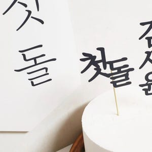 May include: A white cake with black Korean calligraphy cake toppers that say "First Birthday" in Korean. The cake is on a white surface.