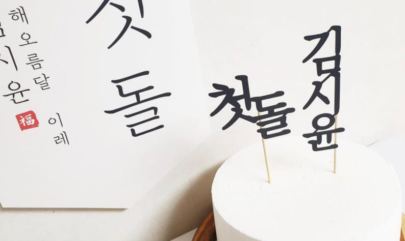 May include: A white cake with black Korean calligraphy cake toppers that say "First Birthday" in Korean. The cake is on a white surface.