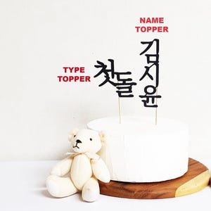 May include: Black cake topper with the Korean text "첫돌" and "김정윤" on a white cake. The cake is on a wooden base. A white teddy bear is sitting in front of the cake.