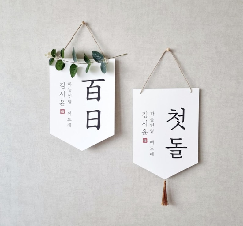 May include: Two white banners with black calligraphy hanging on a wall. The banner on the left says "Hundred Days" in Korean and the banner on the right says "First Day" in Korean.