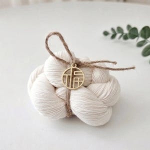 May include: A skein of off-white yarn tied with brown twine, featuring a circular gold-toned metal charm with an intricate cut-out design of the Chinese character for good fortune, known as Fu, centered on the front of the yarn bundle.
