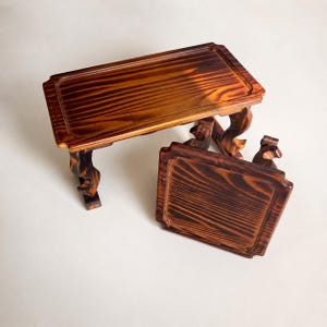 May include: Two small wooden tables with a dark, burnt-wood finish and visible grain patterns. One table is rectangular and standing upright, while the other is square and positioned at an angle, both featuring curved, decorative legs.