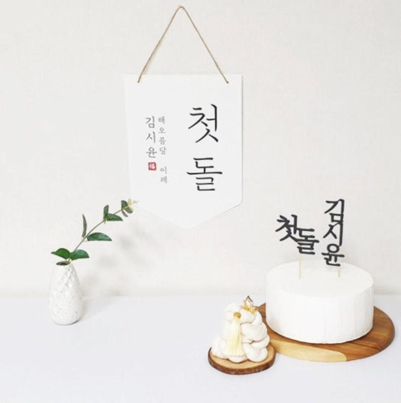 May include: A white cake with black Korean calligraphy on top. The cake is on a wooden cake stand. A white vase with a small green plant is on the left side of the image. A white banner with black Korean calligraphy hangs above the cake. The banner reads "Just Do It".