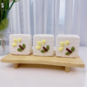May include: Three white square-shaped cakes decorated with yellow and green floral designs on a wooden stand.