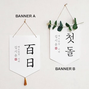 May include: Two white banners with black calligraphy hanging from twine. The banner on the left says "百日" and the banner on the right says "첫돌". Both banners have the text "김해 신문 일례" in smaller font.