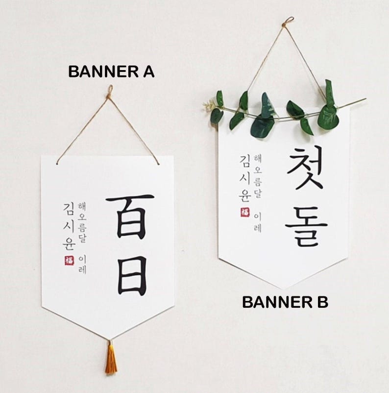 May include: Two white banners with black calligraphy hanging from twine. The banner on the left says "百日" and the banner on the right says "첫돌". Both banners have the text "김해 신문 일례" in smaller font.