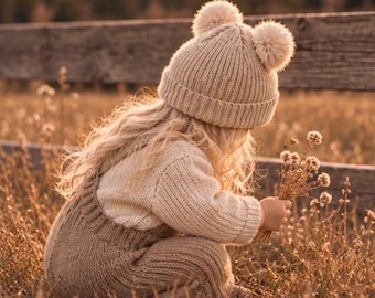 Peut inclure: Un bonnet en tricot beige avec deux pompons sur le dessus, associé à un pull en tricot beige à manches longues et une salopette en tricot assortie. L'ensemble présente une texture côtelée sur toute la surface, créant un look monochrome.