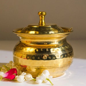 Puede incluir: Un recipiente de metal dorado con tapa y acabado martillado. El recipiente está decorado con una banda de líneas horizontales. El recipiente está sobre una superficie blanca con flores blancas.