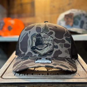 Puede incluir: Gorra de camionero de camuflaje con parte trasera de malla negra y visera curva. La parte delantera presenta un parche con un gráfico de ave volando y el texto "CHUKAR HUNTERZ". La gorra se muestra sobre una superficie de madera.