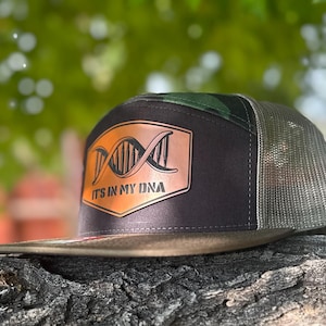 Op de afbeelding: Een zwarte en olijfgroene truckerpet met een mesh achterkant. Het voorpaneel heeft een koperkleurige patch met een DNA-strengontwerp en de tekst "IT'S IN MY DNA". De pet heeft een gebogen klep.