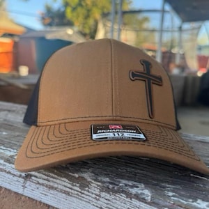 May include: A brown trucker hat with a black mesh back and a stitched brim. The hat features a black cross design and the Richardson 112 logo. Adjustable fit.