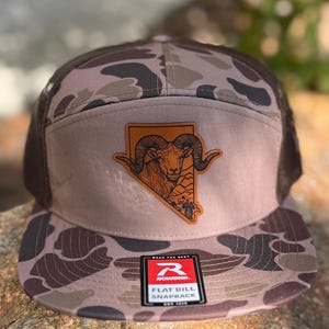 May include: A flat bill snapback hat with a brown and tan camouflage pattern. The hat features a leather patch with a ram illustration and the outline of Nevada. The hat has a Richardson tag.