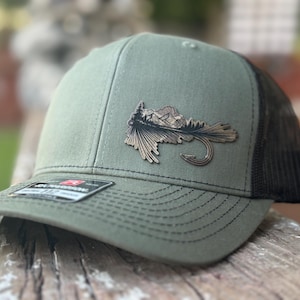 以下が含まれることがあります： オリーブグリーンと黒のトラッカーハット。山と森の風景と釣り針が描かれた、詳細な金属製のエンブレムが付いています。帽子はカーブしたつばとメッシュの背面を備えています。