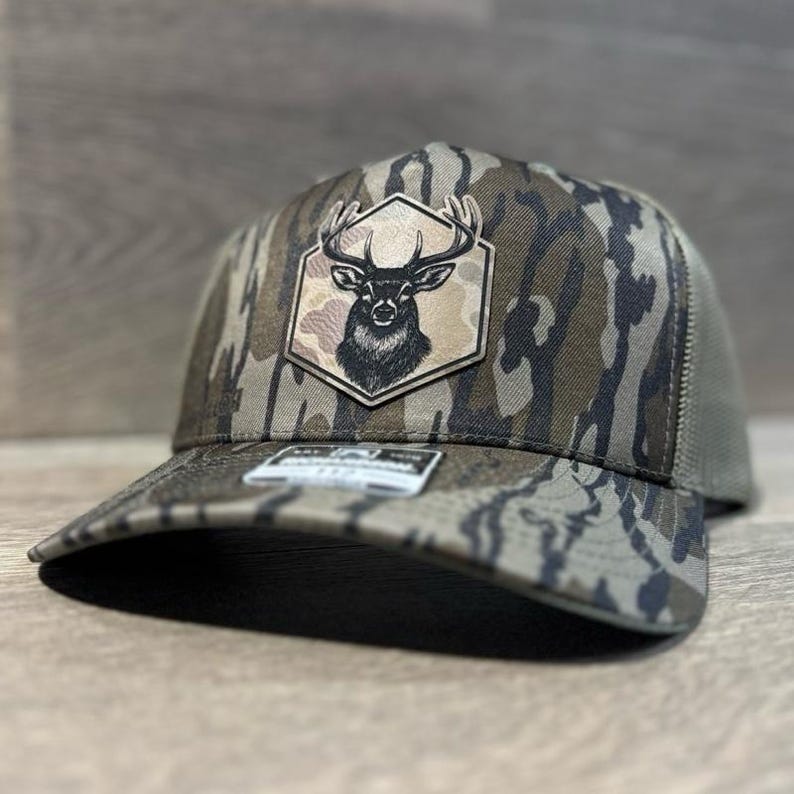 May include: A camouflage trucker hat with a deer emblem. The hat features a brown and green camouflage pattern on the front panels and bill, with a mesh back. The emblem is a black and white illustration of a deer head inside a hexagonal patch.