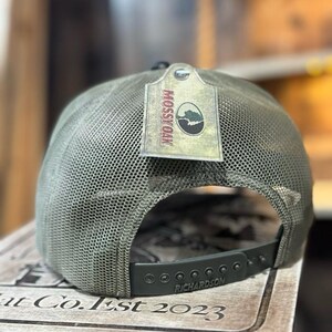 May include: A green trucker hat with a mesh back and a curved brim. The hat features a tag with the "Mossy Oak" logo and the brand name "Richardson" on the adjustable strap. The hat is designed for outdoor activities.