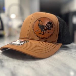 May include: A brown and black trucker hat with a leather patch featuring a rooster and the text "The Frisco".
