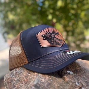 Puede incluir: Gorra de camionero azul marino con la parte trasera de malla marrón y visera curva. La parte delantera presenta un parche color cobre con una ilustración de un alce. El sombrero está sobre una roca.