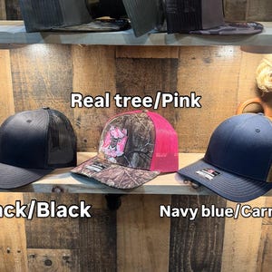 May include: Three baseball caps on a wooden shelf. The caps are black, camouflage with pink mesh, and navy blue with a brown mesh. The text on the image reads "Real tree/Pink", "Black/Black", and "Navy blue/Carmel".