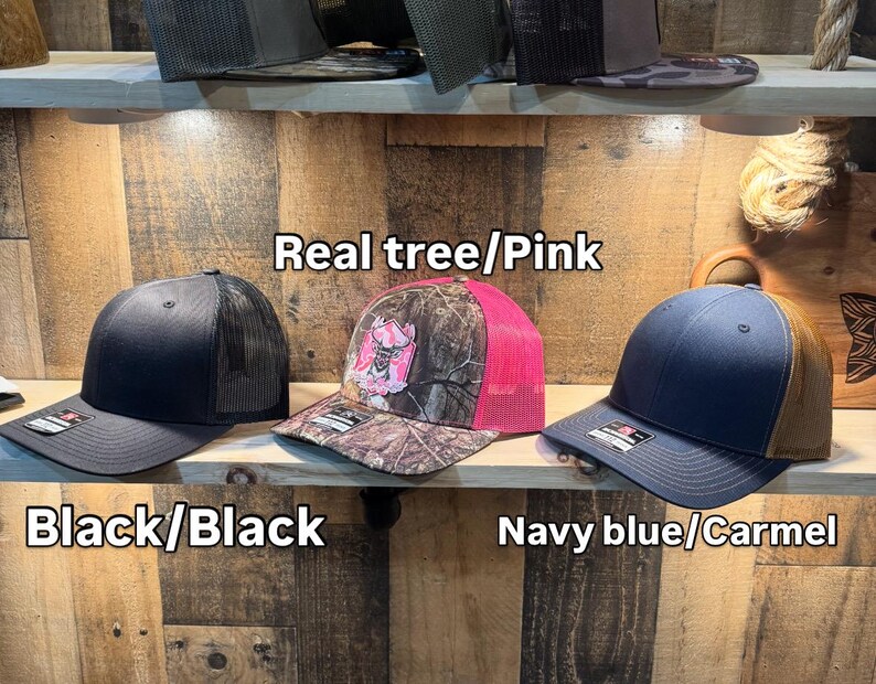 May include: Three baseball caps on a wooden shelf. The caps are black, camouflage with pink mesh, and navy blue with a brown mesh. The text on the image reads "Real tree/Pink", "Black/Black", and "Navy blue/Carmel".