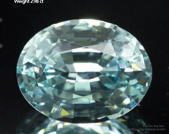 May include: An oval-cut Yttrium Aluminum Garnet gemstone, approximately 9x7mm in size, with a weight of 2.98 carats. The gem has a light blue hue and a faceted surface, reflecting light. The image includes the text "Yttrium Aluminum Garnet" and "GEMTASY".