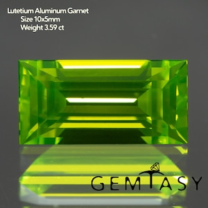以下が含まれることがあります： 長方形の明るい緑色のルテチウムアルミニウムガーネットの宝石。宝石のサイズは10x5mmで、重さは3.59ctです。画像には、下部に「GEMTASY」というテキストが含まれています。宝石は下の表面に反射しています。